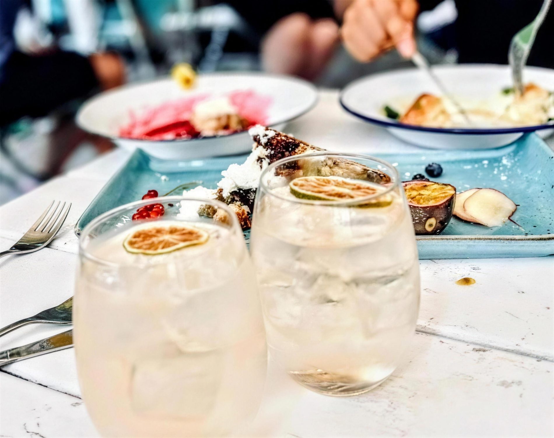 Two iced drinks with dried lime wheels on a white table, brunch plates behind